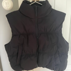 Svart dunväst från H&M Divided - Svart, quiltad dunväst från H&M Divided med hög krage och dragkedja framtill. Västen har två fickor på sidorna och en puffig, oversized look. Perfekt för lager-på-lager-stil under kyliga dagar.