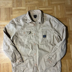 Beige overshirt från Lee i bomull - Snygg beige overshirt från Lee med klassisk krage, två bröstfickor och knäppning framtill. Skjortan är tillverkad i bomull och har diskret fiskbensmönster i tyget. Perfekt lager-på-lager-plagg med avslappnad vibe.