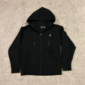 Svart hoodie från Polo Ralph Lauren - Svart hoodie från Polo Ralph Lauren med klassisk huva och snörning. Tröjan har dragkedja framtill, två fickor och den ikoniska broderade loggan i vitt på bröstet. Perfekt för chill dagar och enkel att matcha med jeans eller joggers.