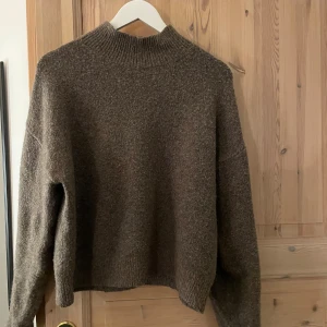 Brun stickad polotröja - Mysig brun stickad polotröja med ribbad hög hals och långärmad passform. Tröjan har en loose och avslappnad siluett, perfekt för att hålla värmen under kyliga dagar. Materialet är mjukt och ger en skön känsla mot huden.