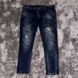 Dsquared2 jeans med färgstänk, stl 52 - Snygga blå jeans från Dsquared2 med coola färgstänk och slitningar på benen. Klassisk femficksmodell med röd patch bak i midjan. Jeansen har en rak passform och är tillverkade i jeansmaterial. Perfekta för dig som vill sticka ut lite extra.
