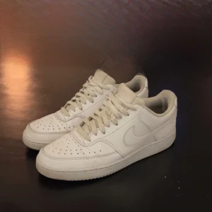Nike Air Force 1  - Klassiska Nike Air Force 1 sneakers i helvitt skinn med perforerad tå, rund tå och platt sula. Snygg och tidlös modell med snörning och diskret Nike-logga på sidan och hälen. Perfekta för dig som gillar en clean och stilren look.
