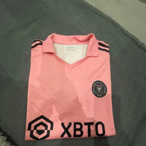 Inter Miami Messi 10 fotbollströja rosa - Snygg rosa Inter Miami fotbollströja från Adidas med Messi och nummer 10 på ryggen. Tröjan har svarta detaljer, klubbmärke på bröstet och sponsortryck framtill. Klassisk V-ringning och korta ärmar, tillverkad i lätt och ventilerande material.