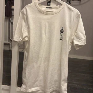 Vit t-shirt från C.P. Company med tryck - Snygg vit t-shirt från C.P. Company i 100% merceriserad bomull. Framsidan har ett litet svart tryck av en person, medan baksidan har ett större tryck med samma motiv och loggan under. Klassisk rund hals och korta ärmar, perfekt för dig som gillar streetwear med stil. Den är i nyskick