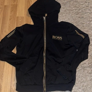 Svart hoodie från Hugo Boss med guld - Svart hoodie från Hugo Boss med guldfärgade detaljer och dragkedja framtill. Tröjan har huva, långa ärmar och snygga guldstreck på axlarna. Boss-logga i guld på bröstet ger en lyxig vibe. Perfekt för dig som gillar stilrena och exklusiva plagg.