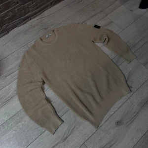 Beige stickad tröja Stone Island - Säljer en beige stickad tröja från Stone Island med klassisk rund hals och ribbade muddar. Tröjan har lång ärm och den ikoniska Stone Island-patchen på vänster arm. Perfekt för dig som gillar clean och stilren streetwear. PASSAR ÄVEN M👍 helt ny aldrig använd ✌️😇