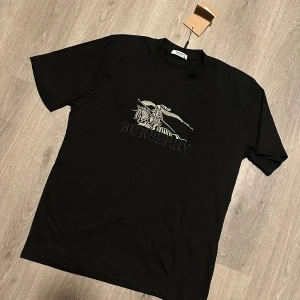 Svart Burberry t-shirt med tryck - Svart t-shirt från Burberry med klassiskt tryck på bröstet i vitt och beige. Modellen har rund hals och korta ärmar, tillverkad i mjuk bomull. Perfekt för dig som gillar stilrena designer med exklusiv känsla.