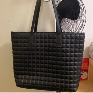 Svart quiltad shoppingväska - Stor och rymlig svart shoppingväska med quiltad design och två långa handtag. Väskan har en fyrkantig form och är tillverkad i skinnimitation som ger en stilren och modern look. Perfekt för att bära med sig allt man behöver under dagen.