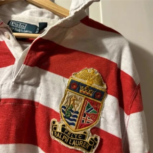 Randig pikétröja från Polo Ralph Lauren - Säljer en klassisk långärmad pikétröja från Polo Ralph Lauren i rött och vitt med breda ränder. Tröjan har en broderad patch med emblem på bröstet och krage med knappar. Perfekt för dig som gillar preppy stil och snygga detaljer.