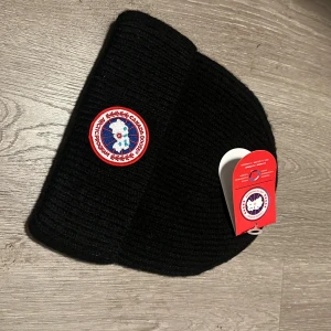 Svart ribbstickad mössa Canada Goose - Svart ribbstickad mössa från Canada Goose med klassisk rund logotyp-patch framtill. Mössan har uppvikt kant och är tillverkad i mjukt, värmande material. Perfekt för kalla dagar och snygg till streetwear-stilen.