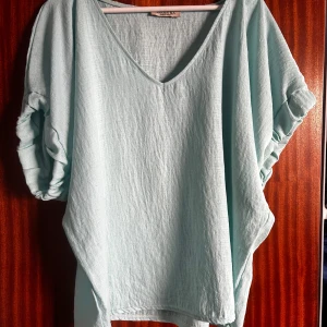 Turkos blus med linnekänsla - TRois KA i Onesize i Viskos och Elastan  Fin vid oanvänd oversized T-shirt med v-ringning och bred resår i ärmsluten (sitter inte åt). Skön viskos i linne-tyg-känsla. Den köpt i butik i Frankrike i somras (för ca 400 kr) men inser nu att det inte är min färg. Personen på bilden är 171 cm lång.  Djurfritt och rökfritt hem.