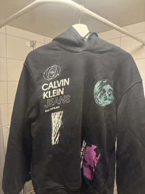 Svart hoodie Calvin Klein Jeans - Svart hoodie från Calvin Klein Jeans med tryck i vitt, turkos och lila på framsidan. Tröjan har huva med snörning och ribbade muddar vid ärmslut och nederkant. Snygg grafisk design och cool streetkänsla.