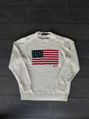 Vit stickad tröja Polo Ralph Lauren - Vit stickad tröja från Polo Ralph Lauren med amerikansk flagga i blått, rött och vitt på bröstet samt RL-broderi. Rund hals och ribbade muddar vid ärmslut och nederkant. Perfekt för dig som gillar klassisk och clean stil.
