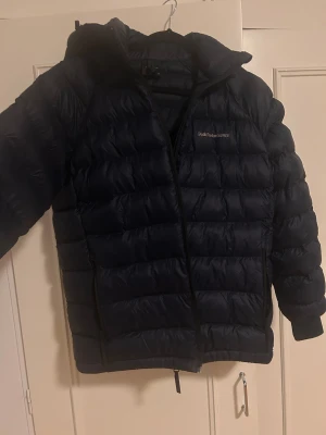 Mörkblå Peak Performance dunjacka - Säljer en mörkblå dunjacka från Peak Performance i storlek 170. Jackan har huva, två sidofickor med dragkedja och quiltad design. Perfekt för kalla dagar och har en clean look med diskret logga på bröstet.