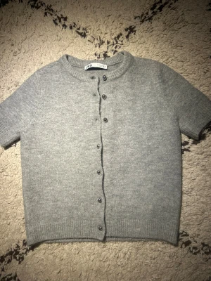 Grå kofta med kort ärm - Grå, mjuk kofta från Zara med korta ärmar och rund hals. Koftan har knappar framtill och ribbade muddar vid ärmslut och nederkant. Perfekt för lager-på-lager och chill stil. Materialet känns mjukt och har en clean look. Ena knappen har tidigare ramlat av men jag har sytt fast den med en knapp på andra sidan (se bild 2). Annars är den i superbra skick. Jag är öppen för prisförslag!🥰