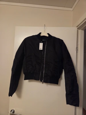 Svart bomberjacka från Gina Tricot - Snygg svart bomberjacka från Gina Tricot med croppad passform och silvrig dragkedja framtill. Jackan har ribbade muddar vid midja och ärmslut, samt klassisk rund krage. Perfekt för dig som gillar en stilren och urban look.