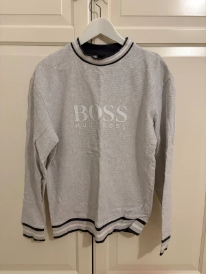 Grå långärmad tröja Hugo Boss - Snygg grå långärmad tröja från Hugo Boss med diskret randigt mönster och vit logotyp framtill. Tröjan har svarta och vita ränder vid hals, ärmslut och nederkant för en sportig vibe. Perfekt för dig som gillar stilrena märkesplagg.