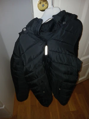Svart pufferjacka med huva - Svart pufferjacka från Explore the Air i storlek 146/152. Jackan har en skön huva, dragkedja framtill och praktiska fickor med knappstängning. Perfekt för kalla dagar och har en stilren look som funkar till det mesta. (Står namn i den)