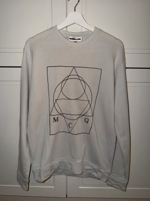 Offwhite sweatshirt från MCQ med tryck - Säljer en clean offwhite färgad sweatshirt från MCQ med ett grafiskt tryck i svart på bröstet. Tröjan har rund hals och långa ärmar, perfekt för dig som gillar minimalistisk stil med en twist. Materialet är mjuk bomull som känns skönt mot huden och är tunn.