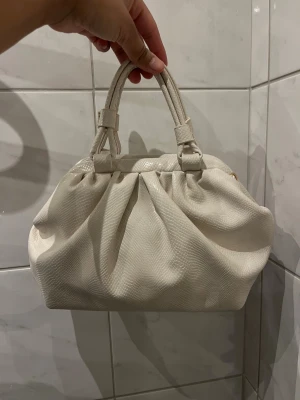 Beige/vit handväska från Fashion Nova - Snygg beige handväska från Fashion Nova med två handtag och ett coolt, lätt glansigt skinnliknande material med diskret struktur. Väskan har en rymlig form med veckad design upptill och guldfärgade detaljer på insidan. Perfekt för dig som gillar stilrena accessoarer.