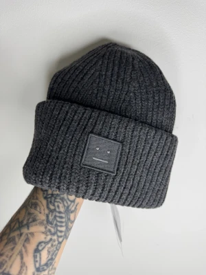 Mörkgrå ribbstickad mössa Acne Studios - Snygg mörkgrå ribbstickad mössa från Acne Studios med ikonisk face-patch framtill. Tillverkad i 100% ull för extra värme och stil. Perfekt för dig som vill ha en clean look med en touch av designer vibes.