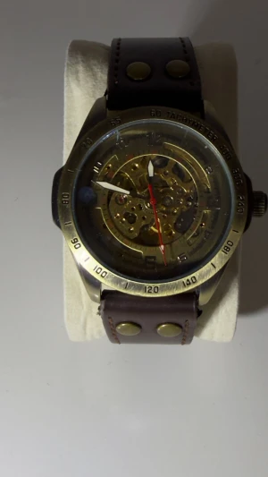 Skelettklocka med brunt läderband - Unik skelettklocka med synligt urverk och detaljerad boett i guldtonad metall. Urtavlan har röda och vita visare samt tachymeter-skala runt kanten. Klockan har ett brett brunt läderband med metallnitar och klassiskt spänne. Perfekt för dig som gillar mekaniska detaljer och cool design.