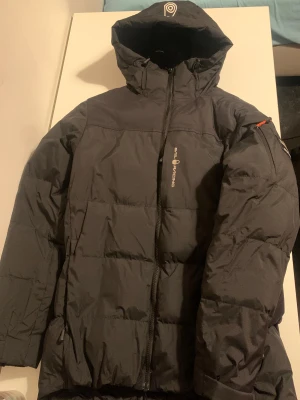 Svart pufferjacka Sail Racing - Svart pufferjacka från Sail Racing med huva och snygga detaljer som patch på ärmen och orange dragkedjedragare. Jackan har en stilren look, lång ärm och logga på bröstet. Perfekt för kalla dagar och expedition vibes.