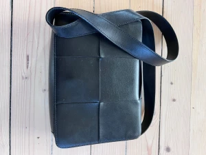 Svart axelväska från & Other Stories - Snygg svart axelväska från & Other Stories i skinn med flätad design på framsidan. Väskan har ett brett axelband och ett innerfack med dragkedja. Perfekt storlek för att få plats med allt viktigt och ger en clean vibe till din outfit. Väskans mått 26x20cm och axelrem ca 1m lång dvs 50cm från axel till väska. Rem kan justeras i 4 olika steg. 