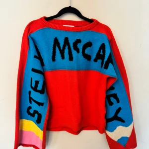 Färgglad stickad tröja Stella McCartney - Säljer en stickad tröja från Stella McCartney med röd bas och blå, gul, rosa och vit färgblock på ärmarna. Svart text med loggan på bröst och ärmar. Tröjan har rund hals och lång ärm. Litet hål längst ut på ena ärmen, se bild.