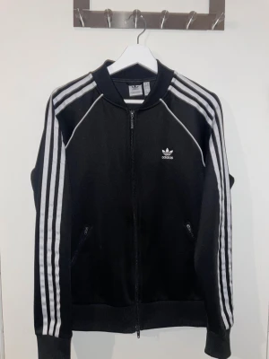 Svart Adidas ziptröja med vita ränder - Säljer en svart Adidas ziptröja med klassiska vita ränder längs ärmarna och Adidas-logga på bröstet. Tröjan har lång ärm, ribbade muddar och dragkedja framtill. Perfekt för en sportig look och chill vardagsstil.