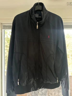 Svart vindjacka från Polo Ralph Lauren - Svart vindjacka från Polo Ralph Lauren med klassisk röd logobrodering på bröstet. Jackan har ståkrage, två fickor med dragkedja och hel dragkedja framtill. Materialet är lätt och syntetiskt, perfekt för blåsiga dagar. Stilren och enkel design som funkar till många outfits.