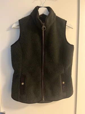 Mörkgrön fleeceväst från Barbour - Mysig mörkgrön fleeceväst från Barbour med hög krage och dragkedja framtill. Västen har två fickor med knappdetaljer och är fodrad på insidan. Perfekt för lager-på-lager och outdoor-vibes.