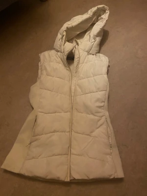 Vit quiltad dunväst från Zara XS - Snygg vit dunväst från Zara i storlek XS. Västen har en quiltad design, dragkedja framtill och en stor huva med snörning. Perfekt för lager-på-lager och chill stil under kyliga dagar. Materialet är mjukt och håller värmen bra.