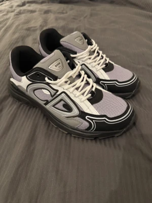 B30 sneakers i svart och grå - Säljer ett par B30 sneakers med chunky sula och coola detaljer. Skorna är svarta, grå och vita med mesh och skinnpaneler, samt tydliga CD-loggor på sidan och hälen. De har snörning och en modern, sportig vibe som sticker ut.