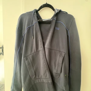 Mörkblå hoodie från Hugo Boss - Säljer en mörkblå hoodie från Hugo Boss med blå detaljer och broderad logga på bröstet. Det finns inga skador, den är inte solblekt och den är i väldigt bra skick. Sällan använd då de inte är min stil. Kom gärna med frågor och funderingar