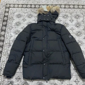 Canada Goose jacka - Svart dunjacka från Canada Goose med pälskant på huvan och klassisk logga på ärmen. Jackan har en quiltad design, dragkedja och knappar framtill samt flera fickor. Perfekt för kalla vinterdagar och har en snygg, tidlös look.