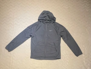 Nike Miller  - Snygg grå vindjacka från Nike med huva och dragkedja framtill. Jackan har två fickor med dragkedja och klassisk Nike-logga på bröstet. Perfekt för löpning eller när du vill ha en lätt jacka med sportig vibe.