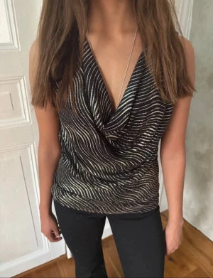 Glittrig svart och guldig topp - Säljer en glittrig topp i svart och guld med vågigt mönster. Toppen kan stylas både med djup v-ringning eller offshoulder. Materialet är mjukt och stretchigt, perfekt för att sticka ut. Passformen är loose och toppen har en snygg drapering framtill.