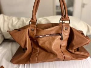 Brun axelväska från Elegance - Snygg och rymlig brun axelväska från Elegance i mjukt skinnmaterial. Väskan har flera praktiska fack med dragkedja och robusta handtag. Perfekt för dig som vill ha en stilren och klassisk look med plats för allt du behöver.