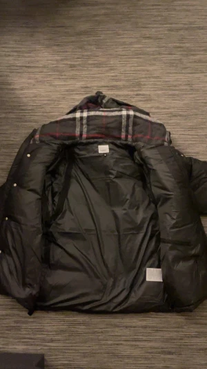 Svart dunjacka från Burberry - Svart pufferjacka från Burberry med stor huva och klassiskt rutigt foder i grått, vitt och rött. Jackan har dragkedja och knappar framtill, samt två sidofickor med dragkedja. Burberry-logga på bröstet och etikett på insidan. Snygg och varm för kalla dagar.