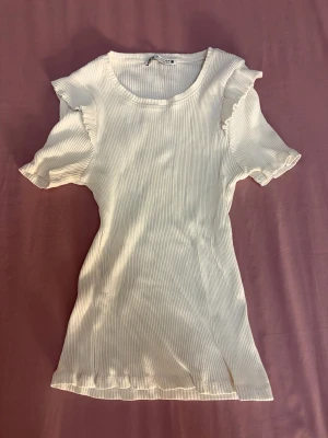Vit ribbad topp med volang från Zara - Supersöt vit topp från Zara!!  I ribbad bomull med korta ärmar och volangdetaljer vid axlarna. Den har en rund halsringning och sitter snyggt med en slim passform! Perfekt för dig!!💕