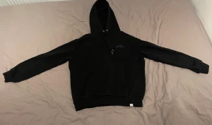 Les Deux svart hoodie - Svart hoodie från Les Deux i storlek S. Tröjan har broderad logga i lila på bröstet, klassisk huva med snörning och en stor magficka. Mjuk och skön bomullskvalitet, perfekt för chill dagar. Hoodien har ett litet hål vid fickan