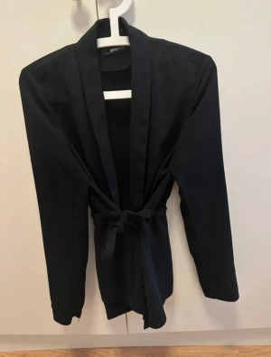 Svart kavaj med knyt från Gina Tricot - Stilren svart kavaj från Gina Tricot med långa ärmar och snygg knytning i midjan. Kavajen har en rak passform och två fickor framtill. Perfekt till jeans eller kostymbyxor för en stilren outfit