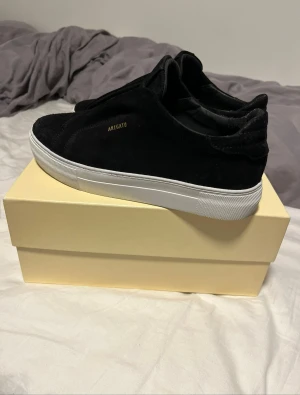 Svarta sneakers från Axel Arigato - Snygga svarta sneakers från Axel Arigato med stilren design och vit platt sula. Skorna är tillverkade i mjuk mocka och har diskret guldfärgad logga på sidan. Perfekta för dig som gillar minimalistisk och modern stil.