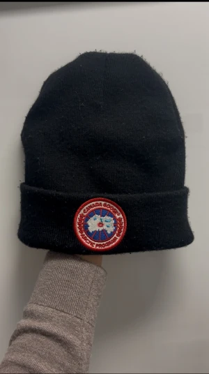 Svart mössa från Canada Goose - Svart ribbstickad mössa från Canada Goose med klassisk rund logopatch framtill i rött, vitt och blått. Mössan har uppvikt kant och liten etikett bak. Perfekt för kalla dagar och har en clean, tidlös look. Riktig 