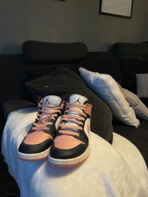 Nike Air Jordan 1 Mid Peach/Black - Nike Air Jordan 1 Mid sneakers i en snygg färgkombo med persikofärgade och svarta paneler, vit bas och matchande persikofärgade snören. Klassisk Jordan-logga på sidan och hög modell som ger extra streetkänsla. Materialet är en mix av skinn och syntet.
