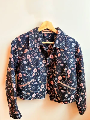 Blommig jeansjacka från Custommade - Säljer en blommig jeansjacka från Custommade med coola detaljer och knappar framtill. Jackan har en mörkblå bas med färgglada blommor i rosa, blått och orange, samt kontrasterande panel på ryggen. Perfekt för dig som vill sticka ut med din stil.