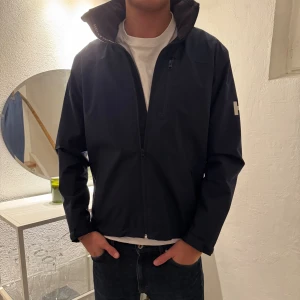 Svart vindjacka från Helly Hansen - Snygg svart vindjacka från Helly Hansen med hög krage och logga på både ärm och rygg. Jackan har dragkedja framtill, flera fickor och är tillverkad i slitstark polyester. Perfekt för blåsiga dagar och enkel att matcha med jeans eller hoodie.