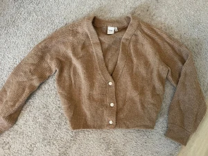 Brun stickad kofta från ICHI - Supermysig stickad kofta från ICHI i storlek L. Koftan har v-ringning, croppad passform och tre pärlknappar framtill. Koftan har en ljusbrun färg
