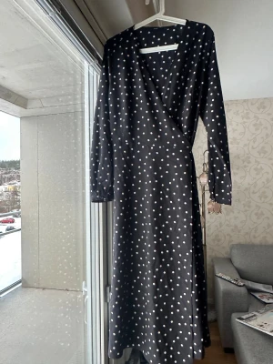 Svart prickig omlottklänning Uniqlo XL - Svart långklänning från Uniqlo med vita prickar, omlottmodell och knyt i midjan. Klänningen har långa ärmar och v-ringad hals. Materialet är mjukt och lätt, perfekt för dig som gillar en flowy look.